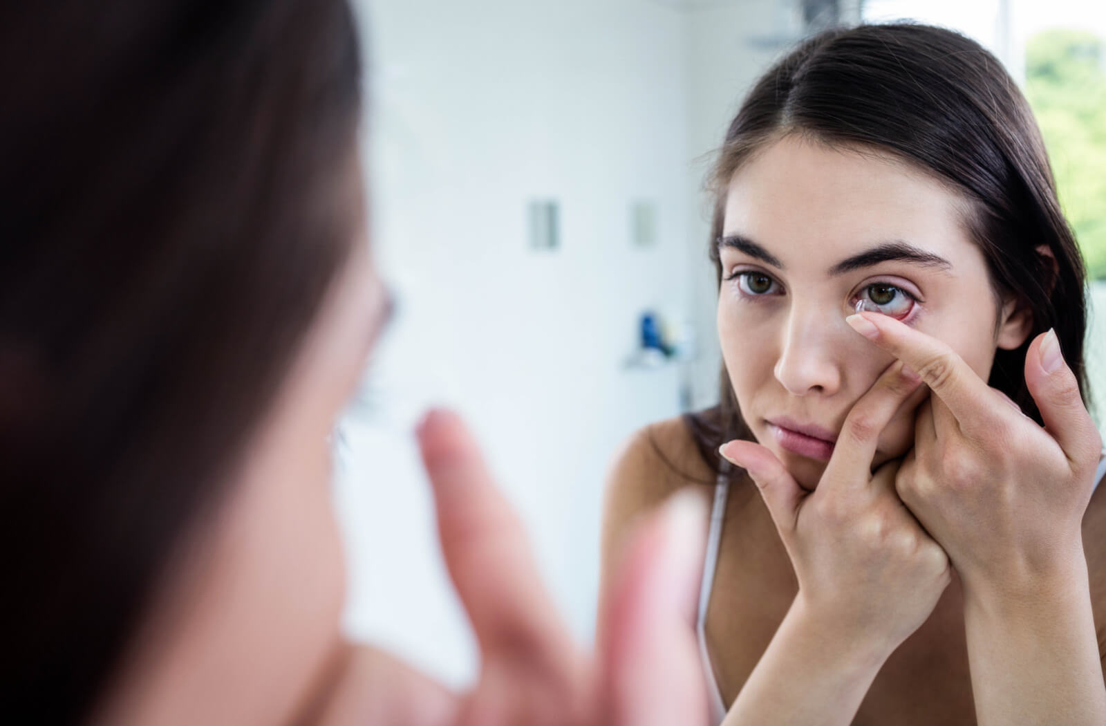A youthful woman with dry eyes is inserting her contact lens while looking into a mirror.