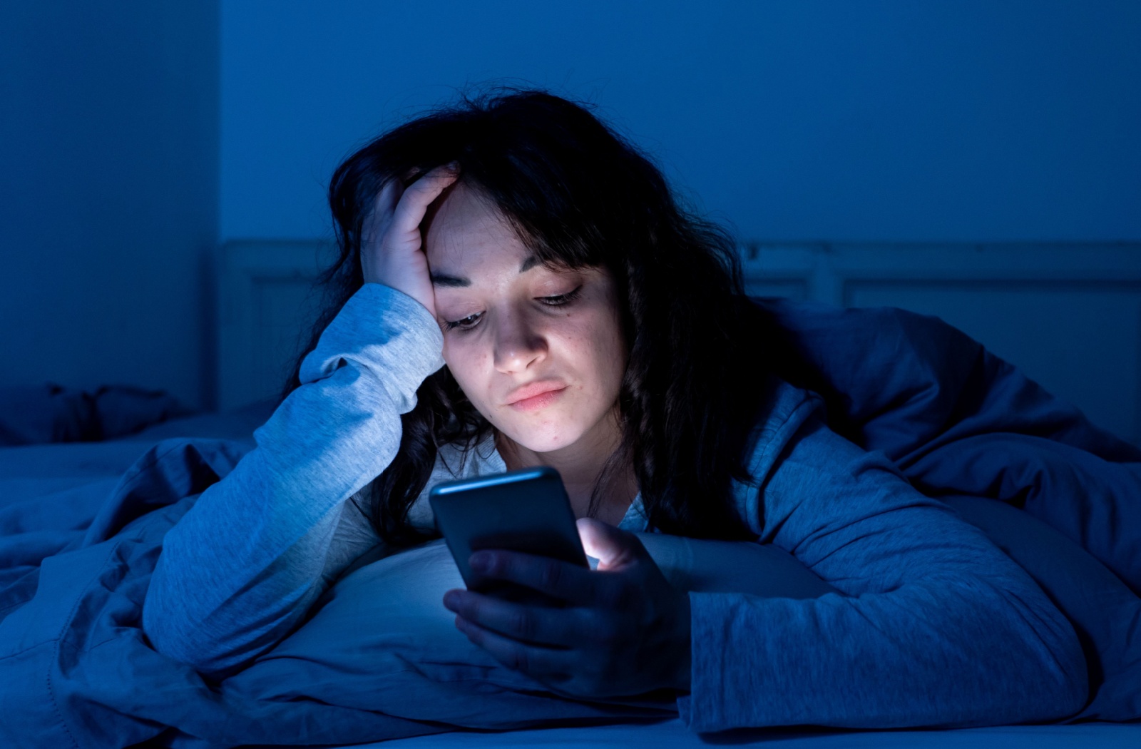A young woman using her cell phone late at night while lounging in her bed. The LED display lights up her face.