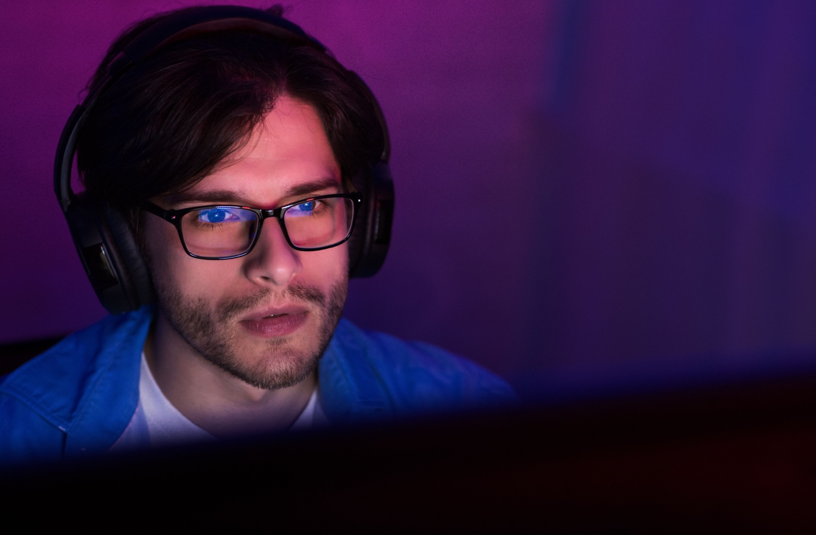 A young mang gaming while wearing blue light blocking glasses, the LED from his computer reflects off the glasses.