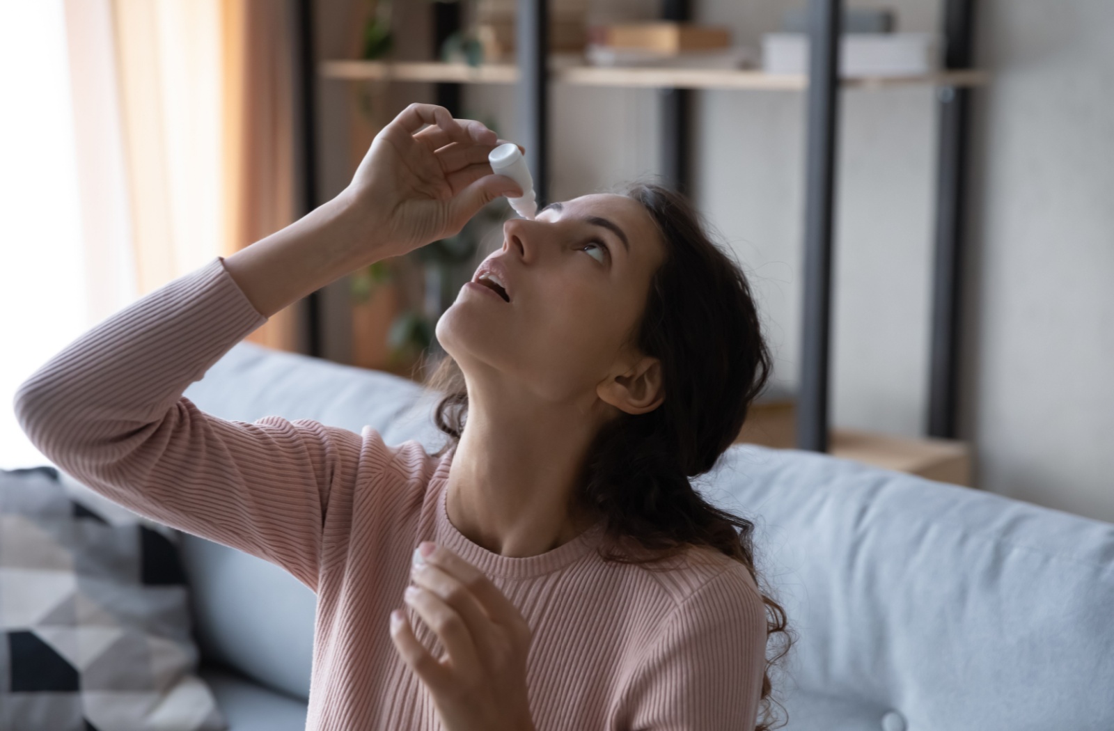 A young woman applies eye drops to find comfort from eye strain.