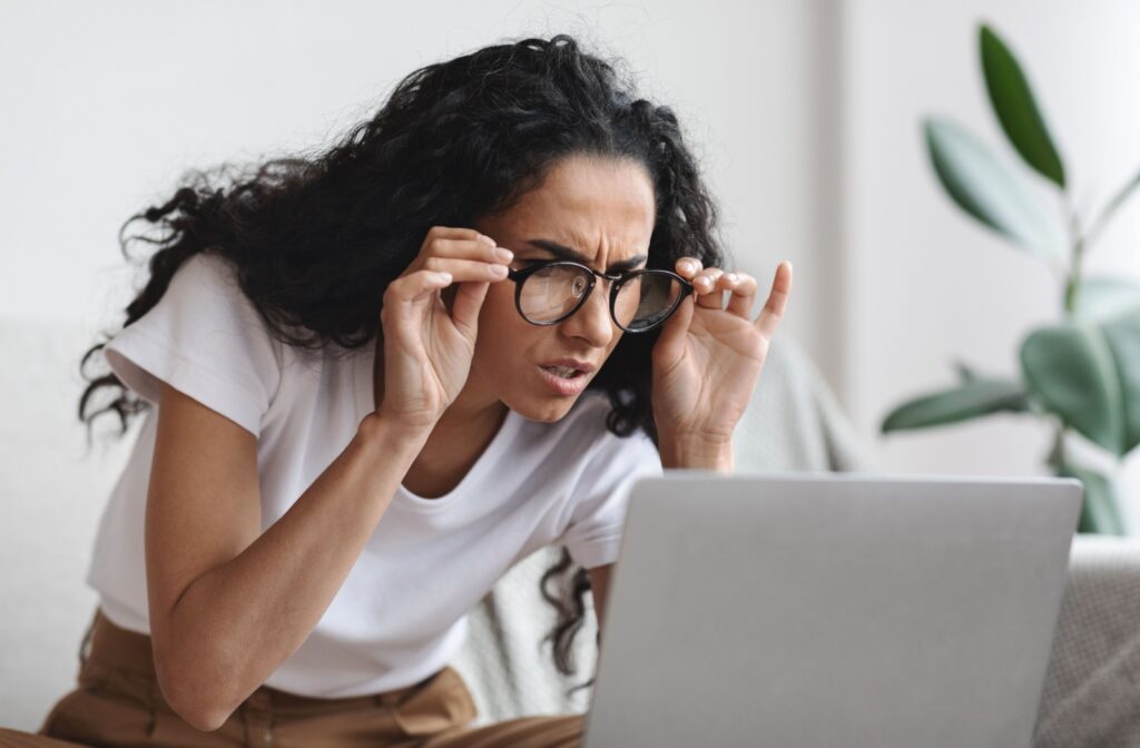 A person squinting to see while leaning over their laptop and adjusting their glasses