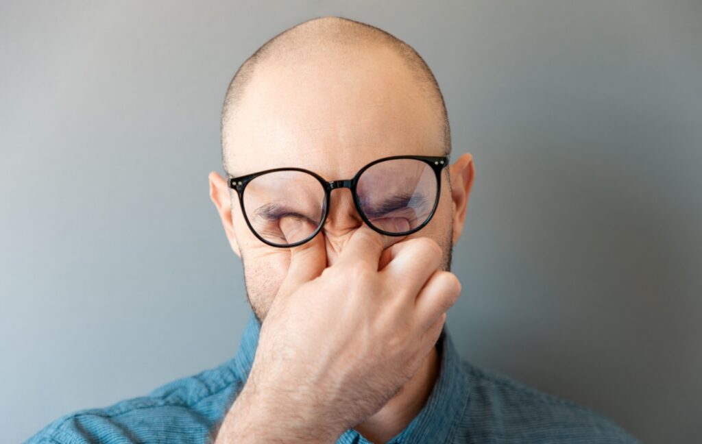 A person lifts up their black-framed glasses to rub their eyes and clear blurry vision caused by dry eyes.