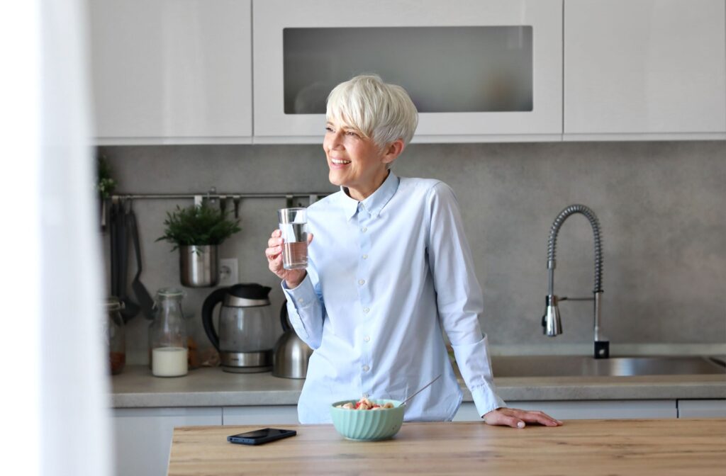An older individual stands in a kitchen smiling as they look out the window while holding a glass of water.