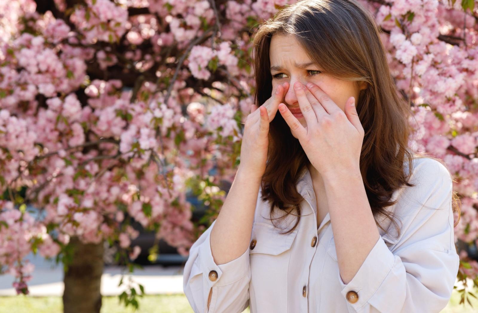 A person rubbing around their irritated eyes while standing outside in front of pink trees in spring