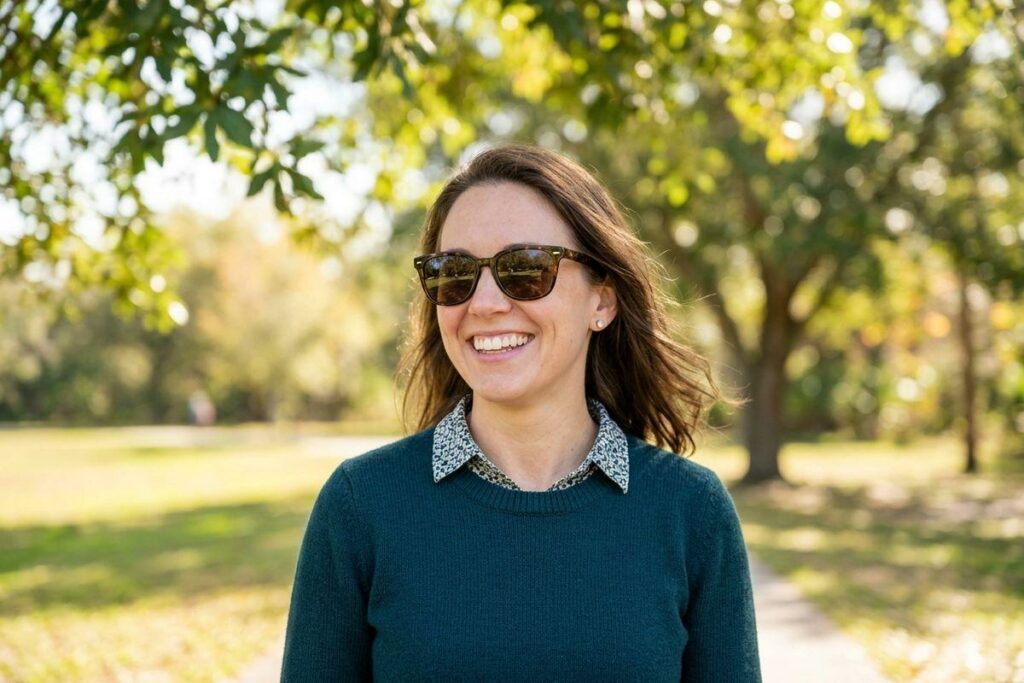 Person in sunglasses and teal sweater smiling outdoors among sunlit trees.